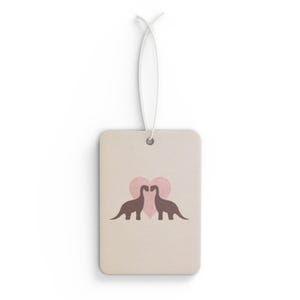 May include: A rectangular air freshener with a light beige background. Two brown dinosaur silhouettes face each other in front of a pink heart. A white string is attached at the top for hanging.