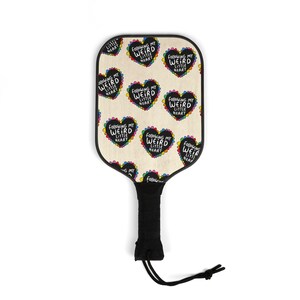 May include: A pickleball paddle with a cream-coloured face featuring black heart graphics with the text "Following My Weird Little Heart" in white. The hearts are outlined with rainbow colours. The handle is black with a black wrist strap.