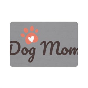 May include: Gray doormat with the text "Dog Mom" in brown cursive lettering. A pink paw print with a white heart inside is above the text.