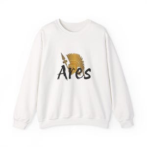 Puede incluir: Sudadera blanca con una ilustración dorada de un casco y una lanza, con la palabra "Ares" en escritura negra. El diseño está centrado en el pecho de la parte superior de manga larga y cuello redondo.
