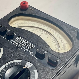 Könnte beinhalten: Ein schwarzes Avometer Analog-Multimeter mit einer großen, gebogenen Skala, mehreren schwarzen Drehreglern und zwei Anschlussklemmen in Rot und Schwarz. Das Gerät enthält aufgedruckte Beschriftungen wie AVOMETER, 6625-99-650-2823 und FOR TRANSIT, SET TO OFF & DC.