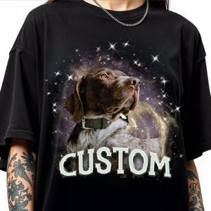 Op de afbeelding: Zwart T-shirt met een hondenportret tegen een ruimte-thema achtergrond, met het woord "CUSTOM" in het wit. De hond heeft bruine en witte vacht en draagt een halsband. Het ontwerp bevat sterren en een nevel.