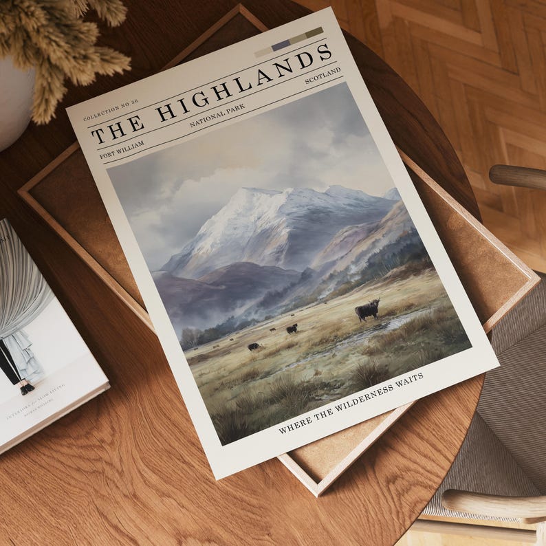 May include: A print titled "The Highlands" depicting a landscape painting of a mountain range with snow-covered peaks, cattle grazing in a field, and the text "Where the Wilderness Waits". The print is on a wooden table.