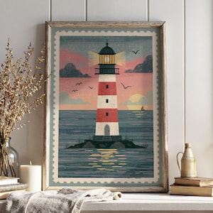 May include: A framed wall art print featuring a stylised illustration of a red and white striped lighthouse on a small island. The scene depicts a sunset over the ocean with birds, clouds, and a sailboat, all framed by a postage stamp border.