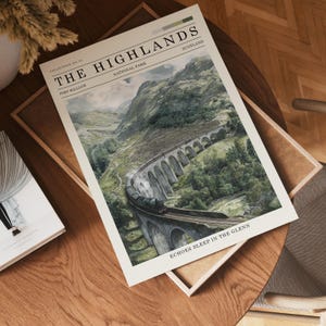 May include: A print depicting the Scottish Highlands, with a train traversing a stone viaduct. The artwork includes the text "The Highlands" and "Echoes Sleep in the Glenn". The print is on a cream background and is displayed on a wooden table.