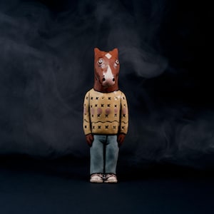 May include: A ceramic figurine of a horse-headed figure wearing a tan sweater with brown and pink accents, blue jeans, and white sneakers. The figure stands against a dark background with a smoky effect.