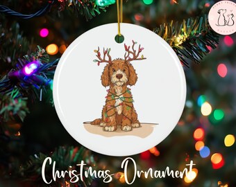 May include: White ceramic Christmas ornament featuring a brown dog with antlers made of Christmas lights. The dog is wrapped in colorful lights. The words "Christmas Ornament" are written in cursive at the bottom.