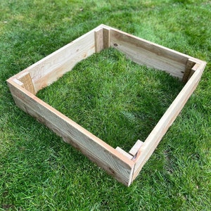 May include: A wooden raised garden bed frame made of untreated wood planks. The frame is sitting on a green lawn.
