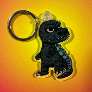 Puede incluir: Un llavero con temática de Godzilla con una ilustración de dibujos animados del famoso monstruo. El llavero tiene un anillo de metal plateado y un accesorio de plástico transparente. La figura de Godzilla es de color verde oscuro con una espina dorsal azul claro y dientes blancos.