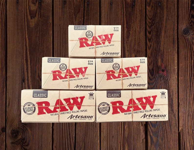 Peut inclure: Une pile de cinq bo&icirc;tes de feuilles &agrave; rouler RAW. Les bo&icirc;tes sont brunes avec un logo RAW rouge et le texte "Artesano" imprim&eacute; dessus. Les bo&icirc;tes sont empil&eacute;es en forme de pyramide sur une surface en bois.