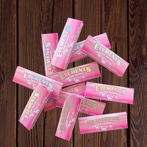 May include: A pile of pink rolling papers with the text "Elements Pink Papers" on each pack. The papers are 1 1/4 size and are vegan.