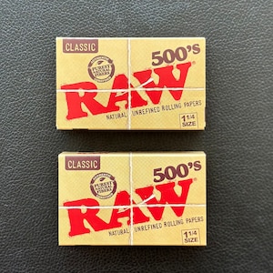 May include: Two packs of RAW Classic 500's natural unrefined rolling papers. Each pack is brown with a red RAW logo and text that reads "Classic", "Authentic", "Pures Natural Fibers", "Unrefined", "500's", "Natural Unrefined Rolling Papers", and "1 1/4 Size".
