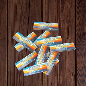 May include: A pile of Elements Ultra Thin Rolling Papers, each pack containing 32 leaves and featuring a vegan gum. The papers are blue and orange with the brand name and details printed on them.