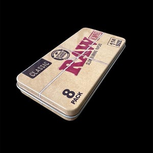 May include: A rectangular, tan metal tin with the word "RAW" in red, and the word "CLASSIC" in black. The tin also has the text "SLOW BURNING PAPERS" and "8 PACK". The tin is 114 size.