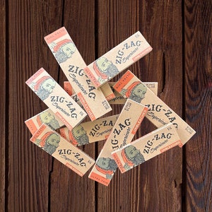 May include: A collection of Zig-Zag rolling paper packs, featuring a bearded man illustration. The packs are beige with red and black text, including "Ultra Thin" and "Unbleached". The papers are scattered on a dark brown wooden surface.