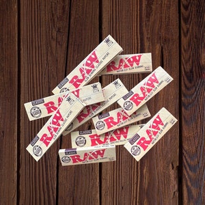 May include: A close-up of a pile of RAW Classic rolling papers. The papers are brown and have the RAW logo in red. The text on the papers reads "SLOW BURNING CLASSIC PAPERS" and "NATURALLY SLOW BURNING".