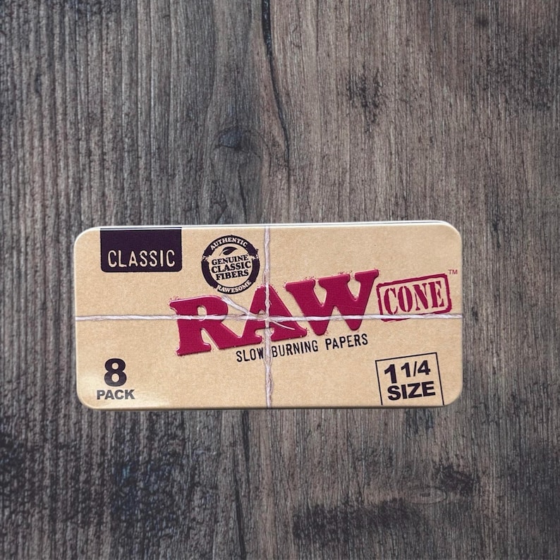 May include: A rectangular box of RAW Cone slow-burning papers. The box is tan with red text that reads "RAW CONE" and "SLOW BURNING PAPERS." It also has the text "8 PACK" and "1 1/4 SIZE." The box is tied with twine.