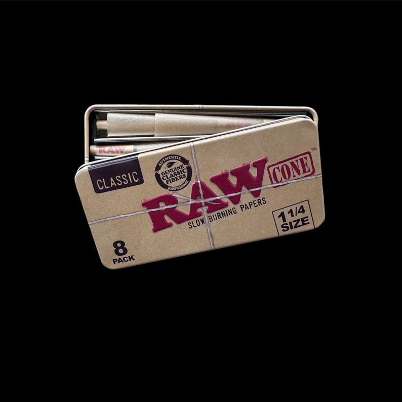May include: A tan metal tin with the word "RAW" in red, and the text "CONE 1 1/4 SIZE" in white. The tin is open, revealing pre-rolled cones. The text "CLASSIC" and "8 PACK" are also visible. The product is designed for slow burning.