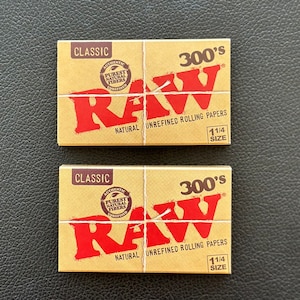 May include: Two packs of RAW Classic 300's natural unrefined rolling papers. Each pack is brown with red text and a red logo. The text on the pack reads "CLASSIC", "AUTHENTIC", "PURE NATURAL FIBERS", "UNREFINED", "300's", "NATURAL UNREFINED ROLLING PAPERS", and "1 1/4 SIZE".