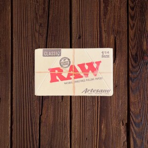 Peut inclure: Un paquet de feuilles &agrave; rouler RAW Classic de taille 1 1/4, non raffin&eacute;es et naturelles. Le paquet est blanc avec du texte rouge et un logo rouge. Le logo dit "RAW" et contient un cercle avec les mots "Authentic Purest Natural Fibers". Le paquet dit &eacute;galement "Artesano Tray Papers Tips".