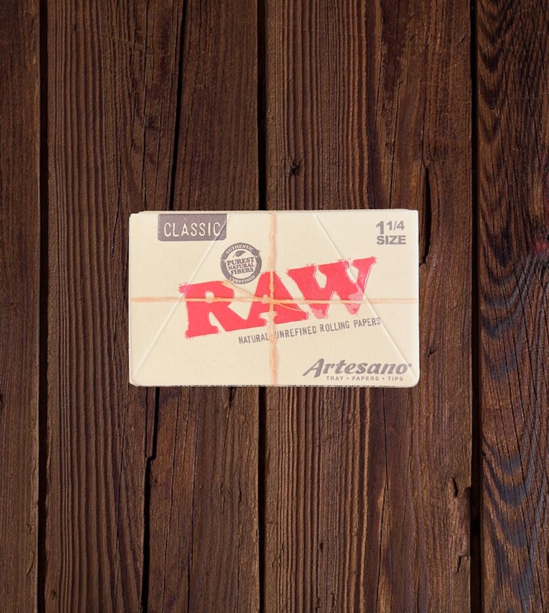 Peut inclure: Un paquet de feuilles &agrave; rouler RAW Classic de taille 1 1/4, non raffin&eacute;es et naturelles. Le paquet est blanc avec du texte rouge et un logo rouge. Le logo dit "RAW" et contient un cercle avec les mots "Authentic Purest Natural Fibers". Le paquet dit &eacute;galement "Artesano Tray Papers Tips".