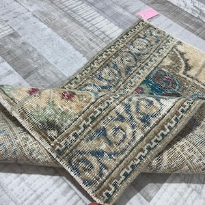 May include: An antique rug with a distressed, vintage appearance. The rug features a beige and brown color palette with accents of blue and red. The design includes floral and geometric patterns, with a detailed border.