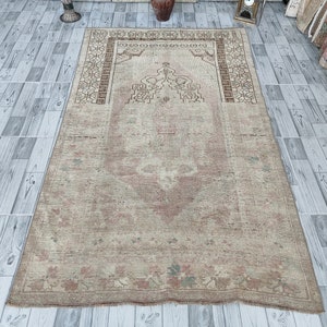 May include: A vintage prayer rug with a beige background and a faded pink and brown floral design. The rug has a central medallion with a geometric pattern and a border with a repeating motif.