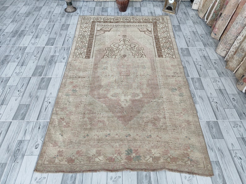 May include: A vintage prayer rug with a beige background and a faded pink and brown floral design. The rug has a central medallion with a geometric pattern and a border with a repeating motif.