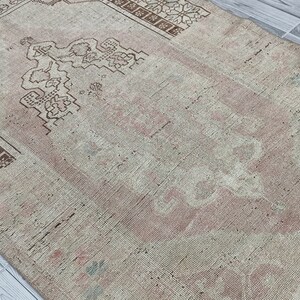 May include: A vintage rug with a faded pink and white color scheme. The rug has a geometric pattern with a central design that resembles a stylized tree.