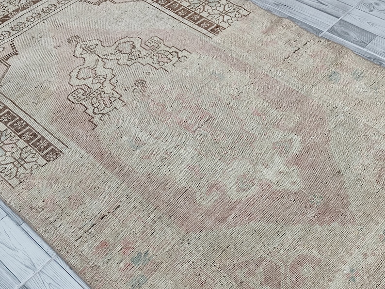 May include: A vintage rug with a faded pink and white color scheme. The rug has a geometric pattern with a central design that resembles a stylized tree.