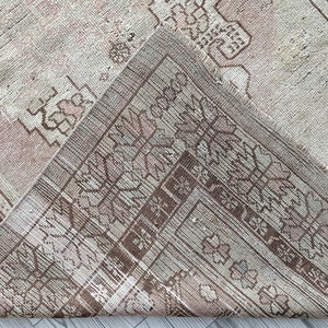 May include: A close-up of a vintage rug with a faded pink and brown geometric pattern. The rug has a worn and distressed look, with frayed edges and a soft texture.