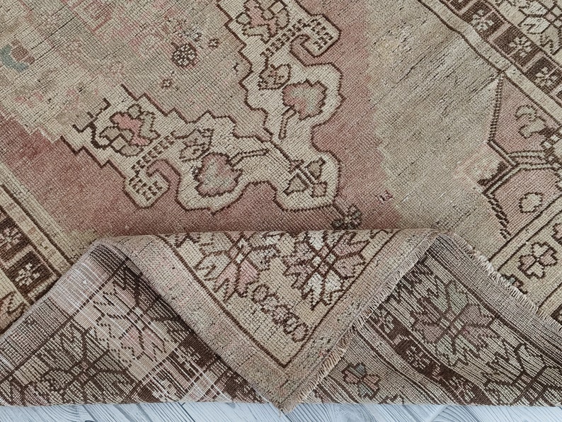 May include: A vintage rug with a light brown and pink color scheme. The rug has a floral pattern with intricate details. The rug is folded in half, showing the underside of the rug.