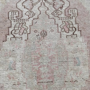 May include: A close-up of a white rug with a faded brown geometric pattern. The rug has a worn and distressed look, with some areas showing signs of wear and tear.