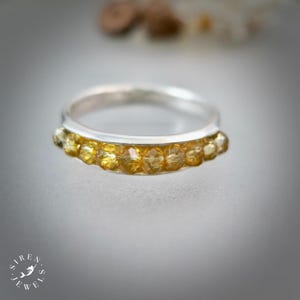 Könnte beinhalten: Ein silberner Ring mit einer Reihe kleiner, facettierter, bernsteinfarbener Kristalle, die entlang der Vorderseite eingelassen sind. Der Ring hat eine polierte metallische Oberfläche und eine glatte, abgerundete Innenschiene. Das Siren Jewels Logo ist unten links sichtbar.