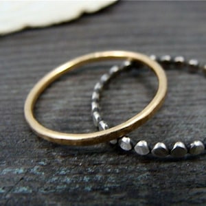 Könnte beinhalten: Zwei Ringe, einer goldfarben und einer silberfarben, werden gezeigt. Der goldfarbene Ring hat eine gehämmert Textur. Der silberfarbene Ring weist kleine, abgerundete Formen auf. Die Ringe sind vor einem dunklen, strukturierten Hintergrund platziert.
