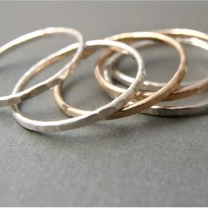 Könnte beinhalten: Eine Ansammlung von gehämmerten Metallringen in Silber- und Goldtönen. Die Ringe sind übereinander gestapelt und zeigen ihre strukturierten Oberflächen und unterschiedlichen metallischen Farbtöne. Die Ringe liegen auf einer grauen Oberfläche.