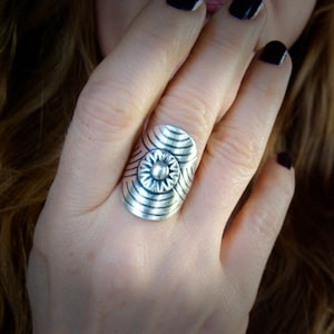 Puede incluir: Un anillo de plata con un diseño geométrico que presenta un motivo floral central. El anillo es ancho y tiene una forma redondeada.