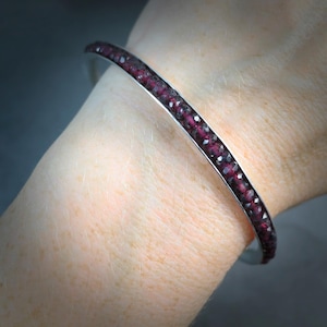 May include: A silver cuff bracelet with a row of small, faceted, deep red gemstones. The bracelet is curved and sits on a wrist, showcasing the jewelry's design and color. The gemstones sparkle, adding a touch of elegance.