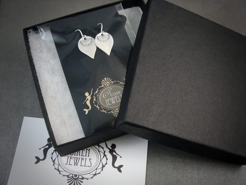 May include: A black box with a white interior, containing two silver earrings with a floral design. The box is open and the earrings are visible. The box has the text "Siren Jewels" printed on it.