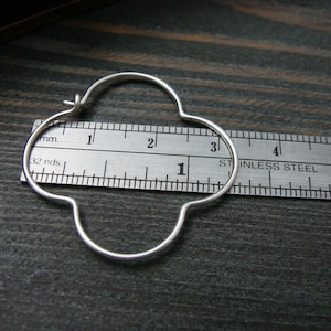 May include: A silver hoop earring with a four-leaf clover design, measured against a ruler. The earring is approximately 1 1/2 inches in diameter.