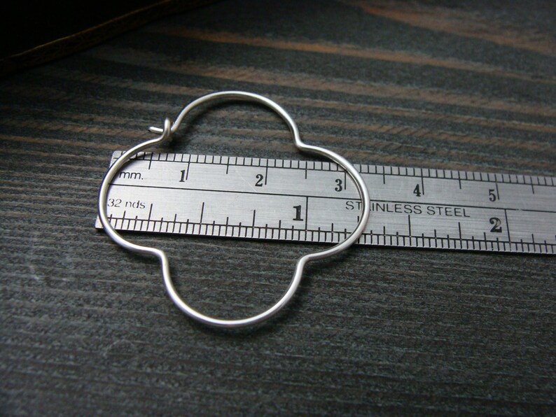 May include: A silver hoop earring with a four-leaf clover design, measured against a ruler. The earring is approximately 1 1/2 inches in diameter.