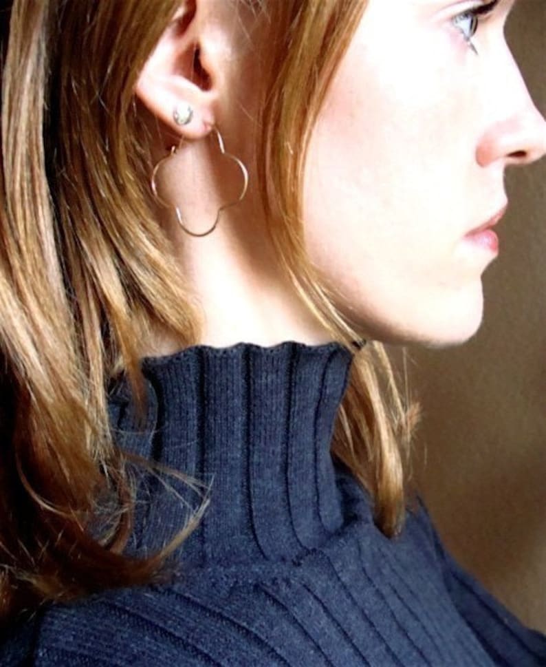May include: A woman with long, straight, reddish-brown hair wearing a black mock neck sweater. She is wearing a pair of gold clover-shaped hoop earrings.