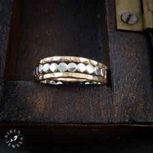 May include: A multi-band ring featuring two outer bands with a hammered gold-tone finish and a central band composed of small, reflective silver-tone circular discs. The ring is positioned within a dark, weathered wooden box with a metal hinge.