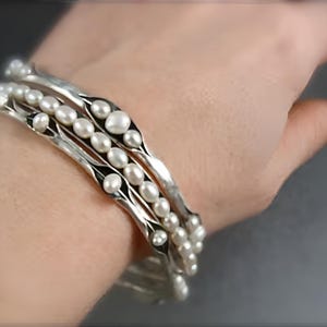 May include: A set of three silver bracelets with white pearls. The bracelets are stacked on top of each other and have a wavy design.