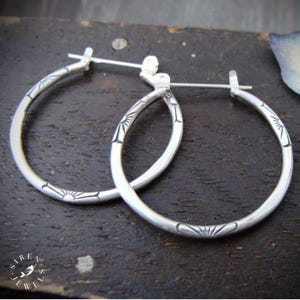 May include: A pair of silver hoop earrings featuring a polished finish and engraved sunburst patterns spaced evenly along the circular bands. The earrings include a hinged post closure mechanism that secures the hoop in a closed position.
