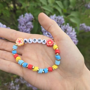 May include: A beaded bracelet featuring flat, circular clay beads in red, yellow, and light blue, interspersed with small gold-toned metallic spheres. The bracelet includes white letter beads spelling ALOHA, flanked by two flower-shaped clay beads.