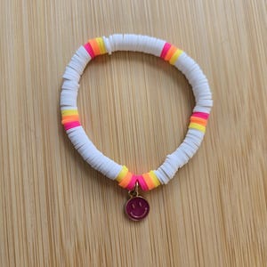 May include: A stretch bracelet composed of small, flat, circular white polymer clay beads interspersed with segments of neon pink, orange, and yellow beads. A small, round, metallic pink charm featuring an engraved smiley face hangs from the center.