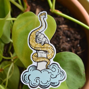 Könnte beinhalten: Ein Aufkleber mit einer gelb-braunen Schlange, die sich um eine Hand windet und aus einer Wolke aufsteigt. Der Schlangenkopf ist erhoben, und die Hand ist weiß mit schwarzen Umrissen. Der Aufkleber befindet sich vor einem Hintergrund aus grünen Blättern und einem braunen Topf.