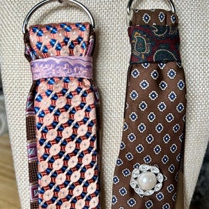 May include: Two keychains crafted from repurposed neckties. The left keychain features a peach and blue geometric pattern with a lavender band. The right keychain has a brown patterned fabric with a central faux pearl and rhinestone embellishment.