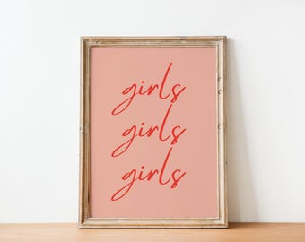 May include: A framed print with the word "girls" repeated three times in a red script font. The background is a solid peach color, and the frame is a light brown wood. The print is displayed on a wooden surface against a white wall.